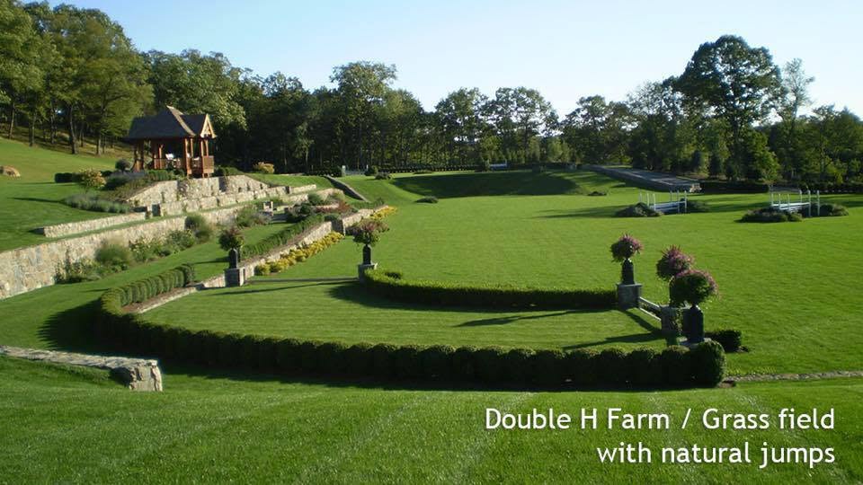 Vue d’ensemble du terrain en herbe de Double H Farm : pavillon, terrasses en pierre, haies taillées et obstacles naturels. Texte intégré : « Double H Farm / Grass field with natural jumps ».